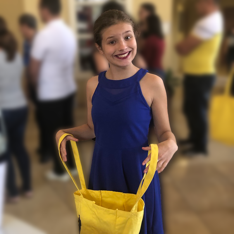 A young girl in a blue dress holding open a yellow bag and looking for donations. She is smiling as she looks at the camera.